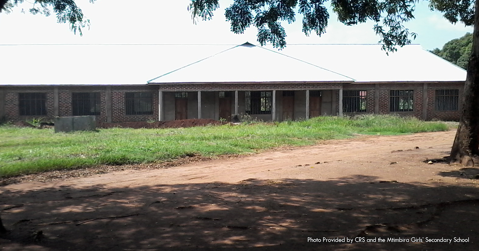 The associated picture was taken and given to us by the Catholic Relief Services. The following picture was taken right outside the Library and Staff room. The Library and Staff room like the rest of the school is made out of bricks. In addition, the top of the Library and Staff room is white in color like the rest of the school. In front of the building are four doors and 7 windows. In front of the 4 doors are 6 gray columns. As for the windows they are all surrounded by the brick layout of the building. In addition, each of the windows have dividers in them that cuts the windows into rectangles. Outside of the building is a small grass plain as well as a dirt road. The grass area has a couple of things that are sitting on top of it. One of the objects that are sitting on the grass is a giant pile of dirt. The pile of dirt is located to the left of the photo and stretches relatively far. The pile of dirt starts from the second door to the left all the way to the last window on the left side of the photo. There is also a gray rectangle block sitting right in front of the pile of dirt. In like manner the dirt road is also in front of the Library and Staff room. The dirt road starts from the front doors of the building and curves outwards toward the picture. Sitting on the side of the dirt road is a giant tree whose leaves provide shade to a section of the dirt road. This building is used to keep books for the student to study. In addition, the building is used by the staff to come to plan out their day.