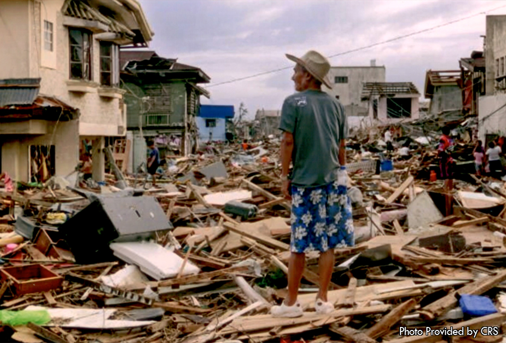 An older gentleman is on looking at the destruction Typhon Haiyan caused in his hometown.  The destruction of the city is so devastating that you can no longer see the road that use to divide both sides of the neighborhood. You can see that there is a huge amount of debris that has fallen from many different places. There are countless amount of pieces of broken wood that have piled up to the point that they create their own hill. Furthermore, there are a lot of pieces of furniture from different buildings and houses that have made their way to the top of the pile of wood. On the left hand side of the photo there are multiple houses who have lost parts of their roof and even parts of their walls. The green house to the left has lost a majority of its roof in the front which was blown into the street. Additionally, the tan house has lost part of its foundation as there is a hole in the bottom left of it. Not to mention, on the right hand side of the photo there are other houses who have lost their roofs and parts of their outside walls.