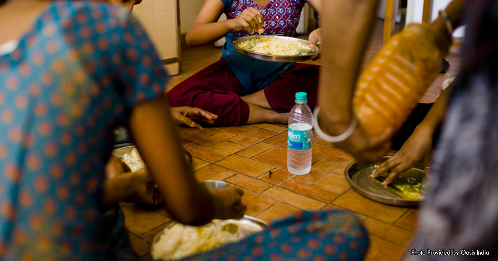 Captured by Tianna, this photo shows a group of 4 girls sitting down on a tiled floor enjoying a meal together. The girls are either survivors or at risk of human trafficking. One way Oasis India helps these young women is by providing housing for them. Although the housing provides the girls with shelter and education, they still need to keep the house in order. The girls living in the house are tasked with a variety of chores to maintain the house. One of these chores is cooking, and the food the girls are eating is a result of them cooking for themselves. On the left side of the photo is one of the girls who is eating her portion of the meal. She is wearing a blue outfit that has a multiple orange spots scattered throughout. To her left is another one of the 4 girls. She to like the rest of the girls is beginning to enjoy her meal. Sitting in the back of the photo is another young girl. With her plate being held in her left hand, she is using her right hand to grab the food off the plate to eat. This young girl is wearing a short sleeved shirt where the front is purple with a patterned design. She is also wearing long pants that are maroon in color. To her left and on the right side of the photo is the last young woman. Unlike the rest of the girls she is sitting with, she is almost down with her food. In the center of the photo, and in the center of the 4 girls is a water bottle. Lastly on the right side of the picture is a man in a black shirt that is holding a container that contains some food for the young girls.