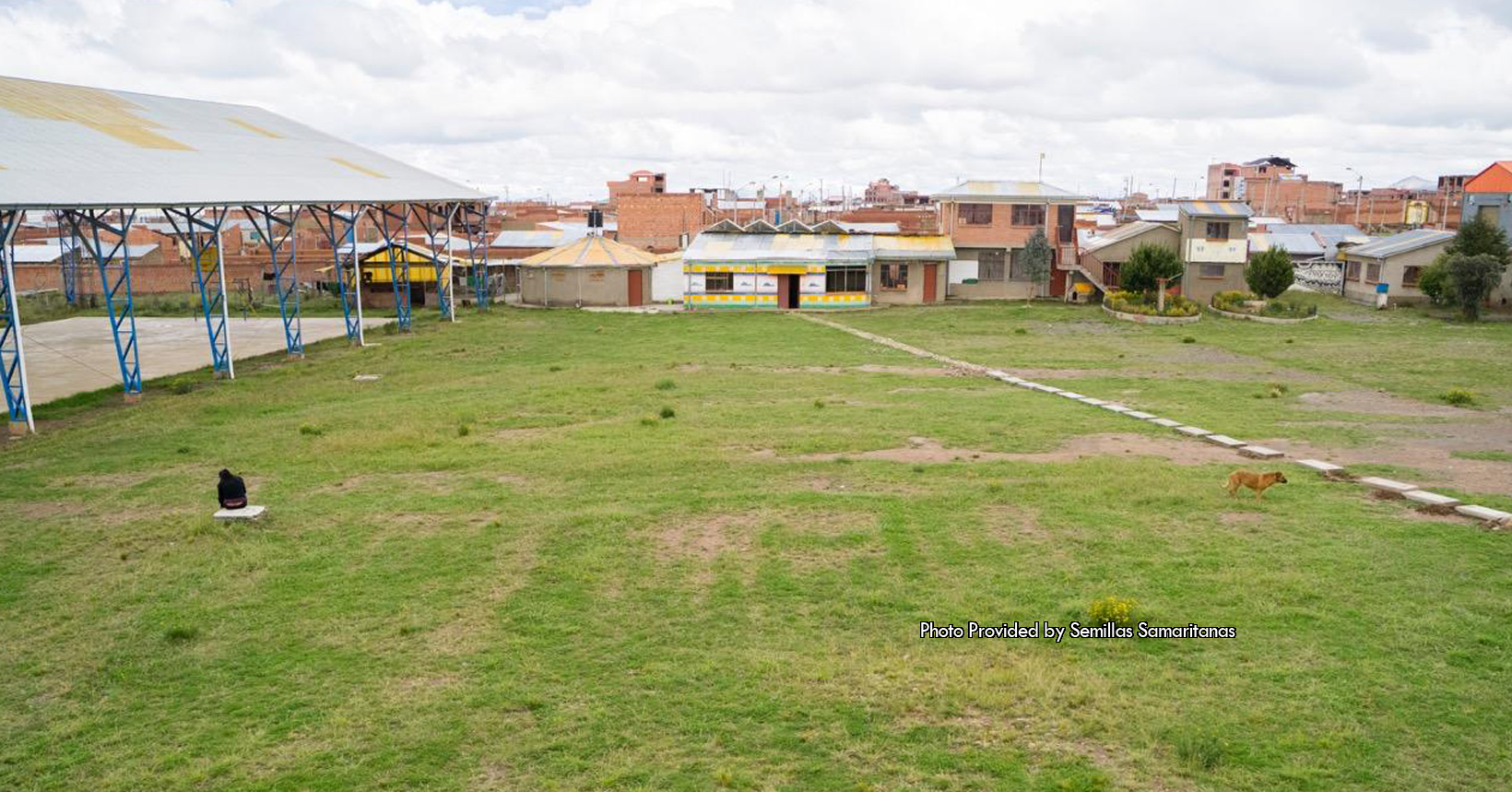 This particular photo was taken outside of the Semillas Samaritanas project. Given to the Father�s Table Foundation, this photo showcases a portion of the futsal court. Futsal is a variant of soccer (or futbol) that is played on a hard court. In addition to being a sector of the futsal court, this area is also a sector of the laundry, bathrooms, bedrooms, study room, television room and computer room. Not to mention, this is also a sector for the housing of all the volunteers. The young kids that come to the Semillas Samartianas project are provided with tools that will ensure their future will be a success. In addition, they are provided a safe place to live for their spiritual and even their psychological recovery. This area is the prime example that does this for the young kids here in the Semillas Samaritanas project. Although this area was and is for the benefit for the kids, they still have to maintain it and keep it clean. In the sector there is an open field of grass that connects multiple buildings together. The field of grass is bright green, but in some areas the grass is slowly dying. Furthermore, to the left of the photo is a concrete area that has a roof over it. The roof is mostly white, but some of the shingles are yellow in color. In the background of the photo there are multiple buildings that the Semillas Samaritanas project uses for various reasons. One of the buildings in the background has four parts of its wall in white. This building has a stone path that stretches from one of its doors all the way to the bottom right hand corner of the photo. Not to mention in the photo there is a person sitting down on a stone tablet and a dog that is standing next
