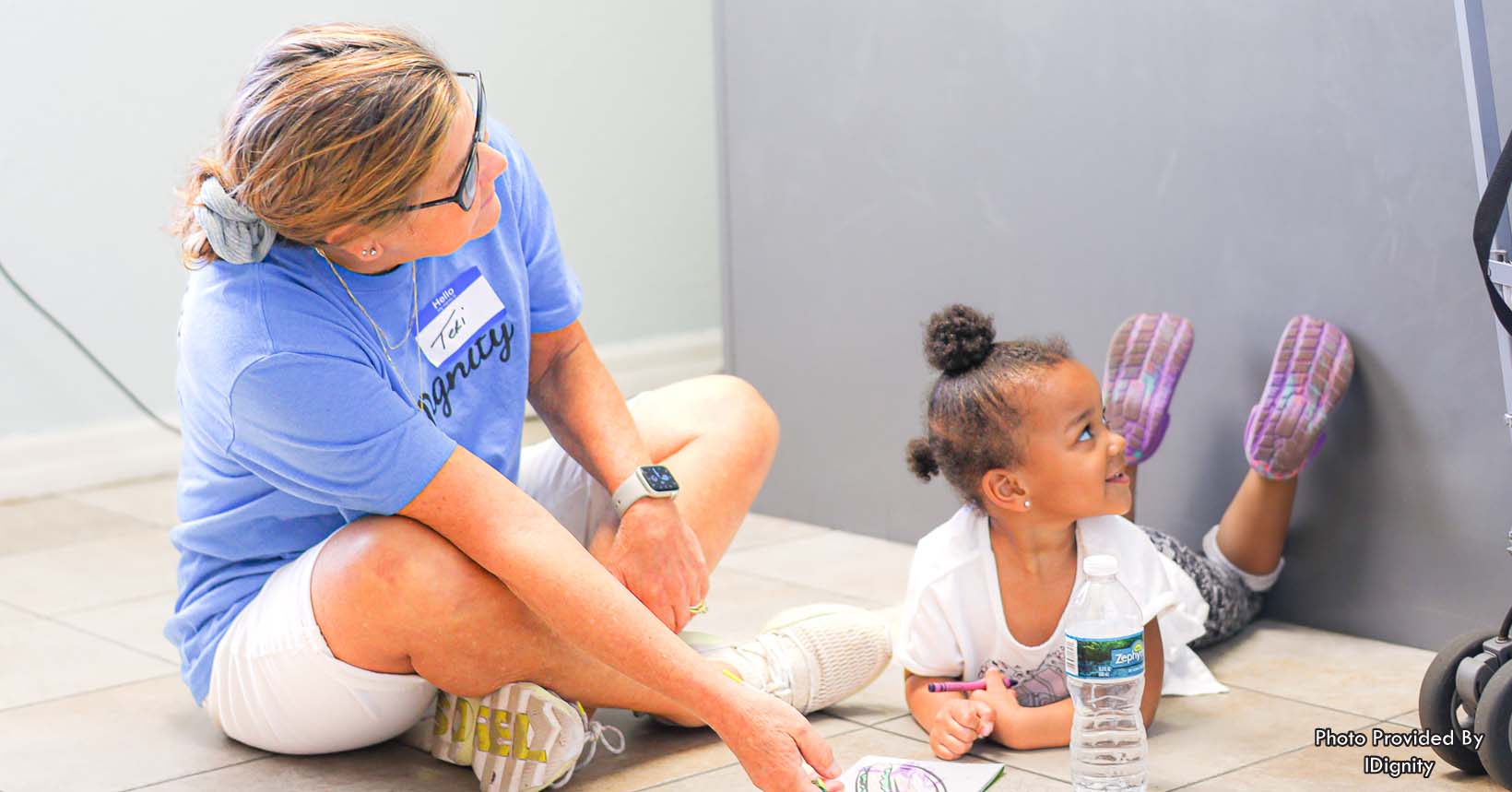 Volunteer Teri sits on the floor with the granddaughter of a client as they work through the process of obtaining her social security card and birth certificate. With these documents, the client and her granddaughter will be able to move out of a shelter into more permanent housing.