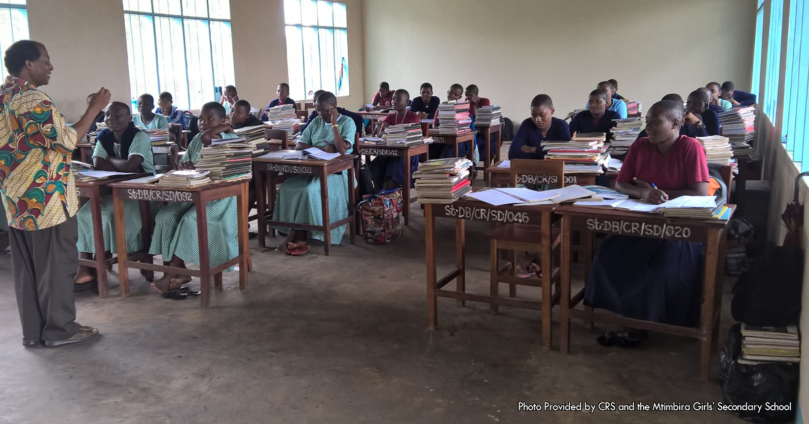 This associated picture was taken by the Catholic Relief Services inside one of the many classrooms. The girls in the classroom are sitting down in their desk preparing themselves for the rest of their lives. The girls in the picture are wearing a variety of clothing but some of them are wearing the same thing. For starters there are a couple of girls who are wearing teal dresses. Three of the 5 girls wearing the teal dresses are sitting in front of the teacher. As for another group of girls, some of them are wearing maroon polo shirts with a blue dress. For the last group of girls they are wearing dresses as well but they are also wearing a dark blue sweater. On top of every desk that the girls are sitting on have multiple books and papers. These are the textbooks that the girls are using to educate themselves as well as notes they have taken and are taking. In addition, written on each of the girl’s desk there are multiple letters and numbers used for labelling. Each desk has the following letters “St-DB/CR/SD” followed by different numbers depending on the desk. Furthermore, all of the girls sitting in the classroom are wearing sandals, some of them being black in color while others are multi-colored. Standing in front of the girls is the teacher for this classroom. He is wearing a shirt with leaves scattered throughout. In addition, the teacher is wearing brown khaki pants and brown sandals. The classroom the girls are sitting in is part of the Mahenge School used to educate the girls. The classroom is white in color, with the floor being gray in color. In addition, the windows of the classroom are blue when you look at them from the side, but are clear looking through them.