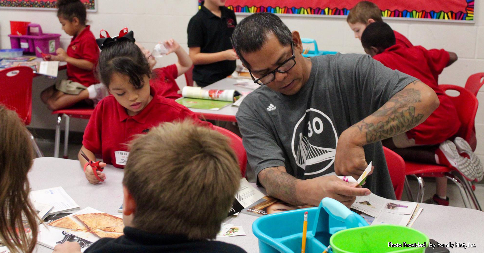 A man in a grey shirt and his daughter sit in her elementary school classroom and work on a crafting project.