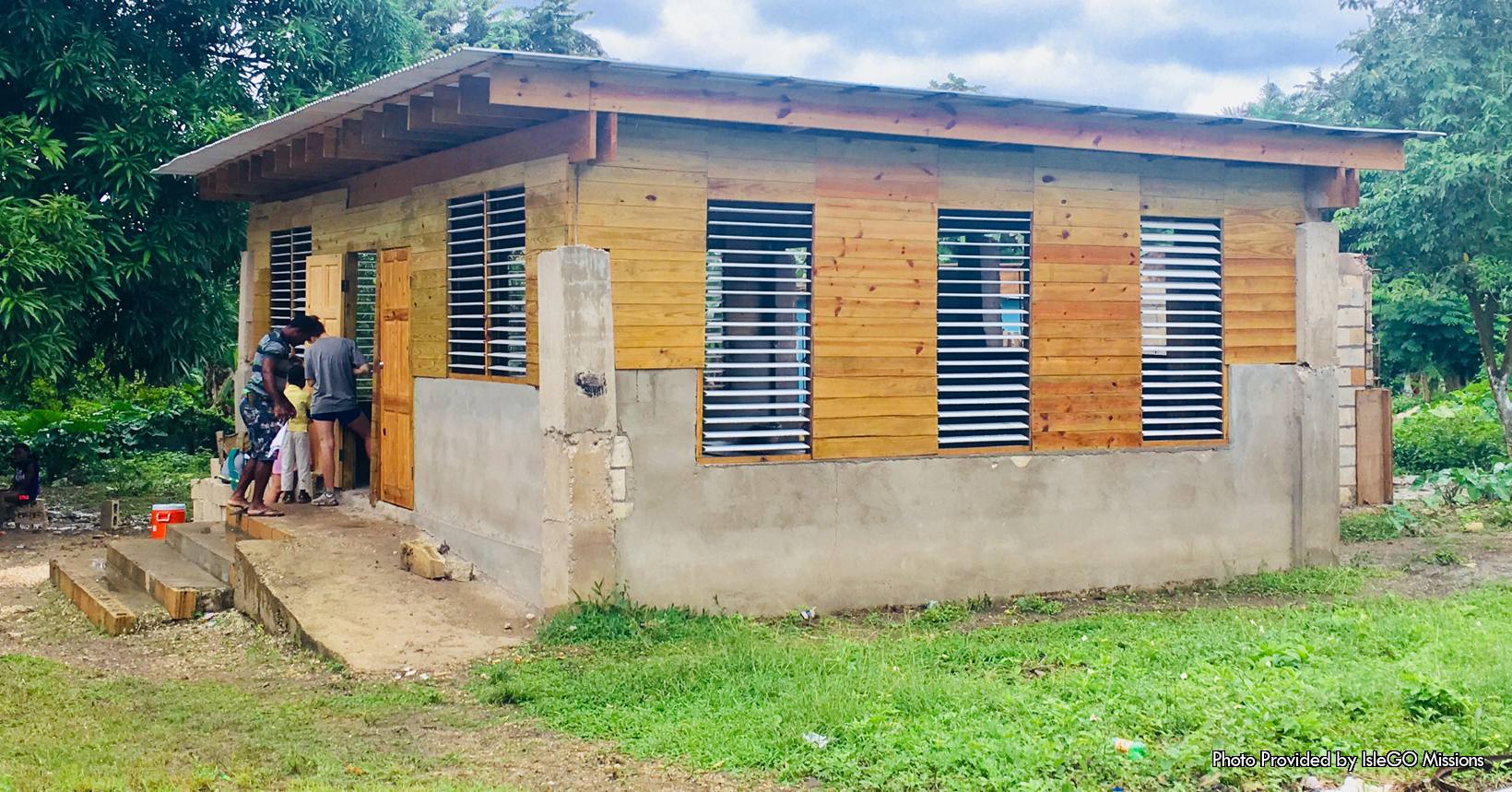 In Jamaica there are many places that are underdeveloped and need much assistance from outside sources. This picture which was taken in Jamaica by IsleGo Missions, illustrates one of the many things that are being developed to help those in need. This picture captures the first Hope Center in Jamaica. The center has four flights of stair that start from the ground up to the front doors. The front doors as well as most of the center is made up of wood. The wood surrounds the Hope Center starting from its half-way point all the way to the top. Furthermore, the wood was placed horizontally around the house. Although the middle and upper-half of the Hope Center is made out of wood, the bottom half is made out of cement. Not only is the bottom half made out of cement, the pillars on the four corners of the Hope Center are also made out of cement. Not to mention, the Hope Center has multiple windows embedded in it. There are two in the front entrance as well as a several around the building. This Hope Center was built in a squatter community known as “the Compound”. The squatter community has around 1500-2500 people in it and they have been forgotten by the Jamaica government as well as the general society. Here in the Compound many suffer from hunger, violence, illiteracy, unemployment, and poverty. In addition, the children in this community suffer from sexual abuse and sometimes the abuse can be from incest. The goal of the center will be to provide jobs for employment, nutrition for the kids, after school programs and last but not least a daycare for parents who are working. In more detail, nutritional meals have already begun to be made for the kids as well as land adjacent from the center being cleared for farming purposes. In the end this Hope Center will be the foundation of hope for the people in this community.