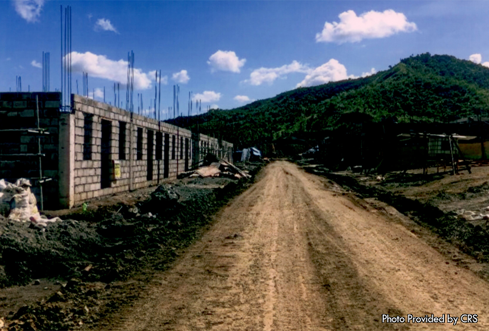 A dirt road bisects the photo with a row of homes in construction.  The houses are build of concrete block and rebar, and awaiting their second floor.