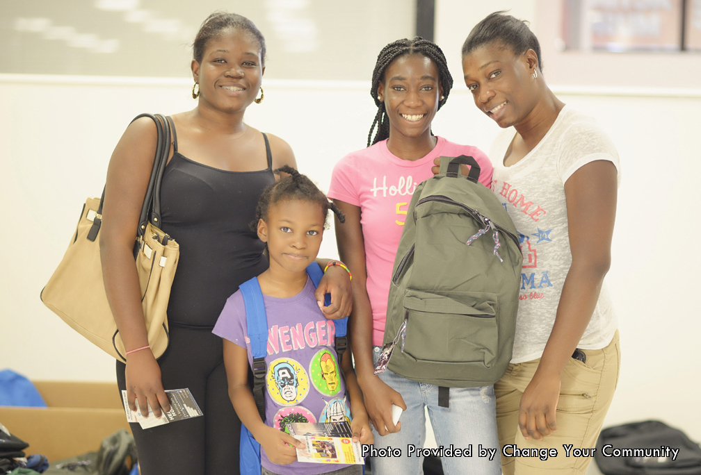 The associated photo was also taken at the same annual Give-A-Way hosted by the Change Your Community Organization. The photo was the given to the Father's table Foundation after the event was over. This photo shows what the Change Your Community mission is all about. In the photo a family is posing after receiving backpacks and school supplies from the Give-A-Way. They can now rest knowing that they don’t have to spend money on school supplies and can use it for something else. In addition, this will mean that the young kids in the family will not have to worry about not having the necessary school supplies. They won’t have to ask their friends or teachers at school to borrow supplies. Starting from the left side of the photo is the mother of the family. She is wearing a black top and black legging pants. She is smiling knowing that her kids are ready to go to school with a backpack and school supplies. Standing to her left is the young daughter of the family. She is wearing a purple shirt that has the Marvels Avengers on it. In her hand is a pamphlet that she received at the event. On her back is her new blue backpack that she is proudly wearing for the first time. To her left are the last two ladies in the photo. The girl in the pink Hollister shirt and light blue jeans is smiling as she holds her new backpack. She too like the rest of the kids who came to the event was given a backpack that will satisfy all of her school needs. Standing next to her is the last lady in the photo. She is wearing a white short sleeved shirt with brown khaki long pants. All of the ladies here has had a problem lifted from their.