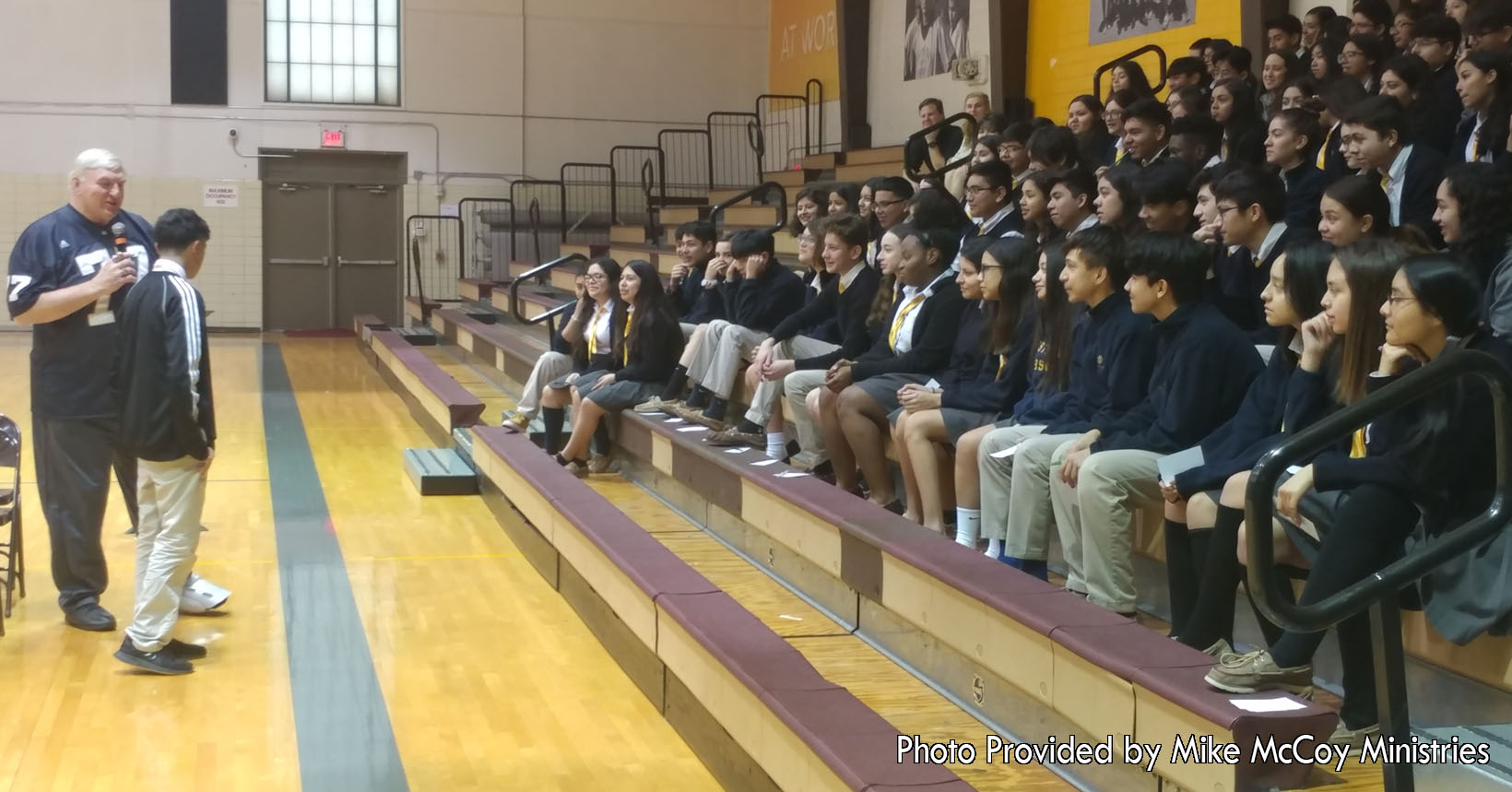 Mike McCoy is giving his faith-based message to a group of young students in one of his many assemblies. The young man standing next to Mike McCoy is participating in the assembly to help the other students get a better understanding.  After this, the students will be given lessons that will help them further in life.