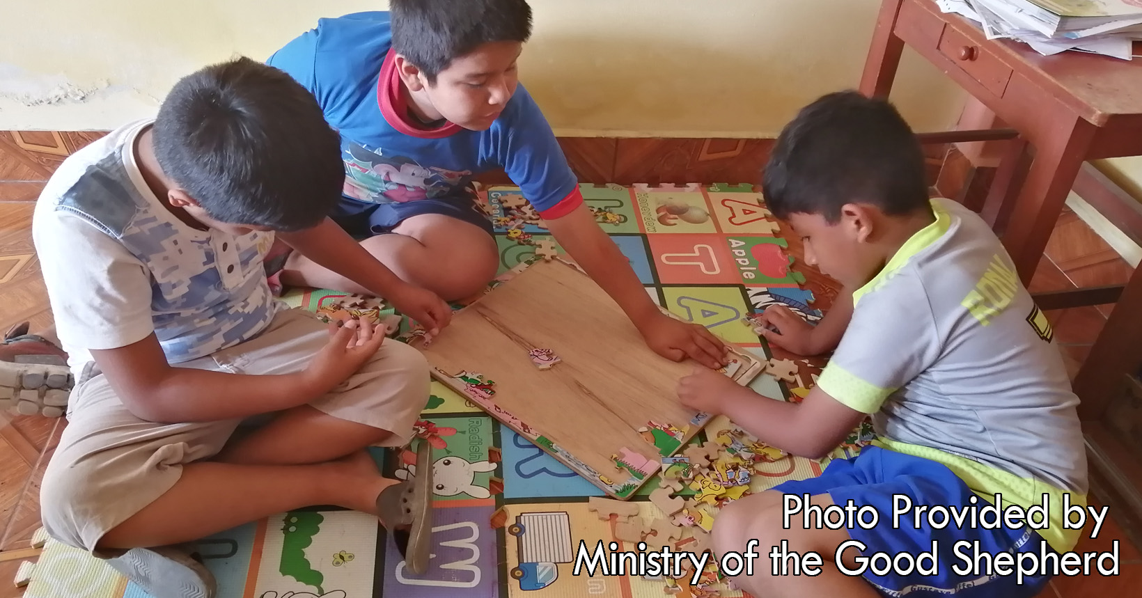 A group of three young boys are playing together in one of the Child Care Centers. They sit upon the floor and are discussing how to put together a puzzle.