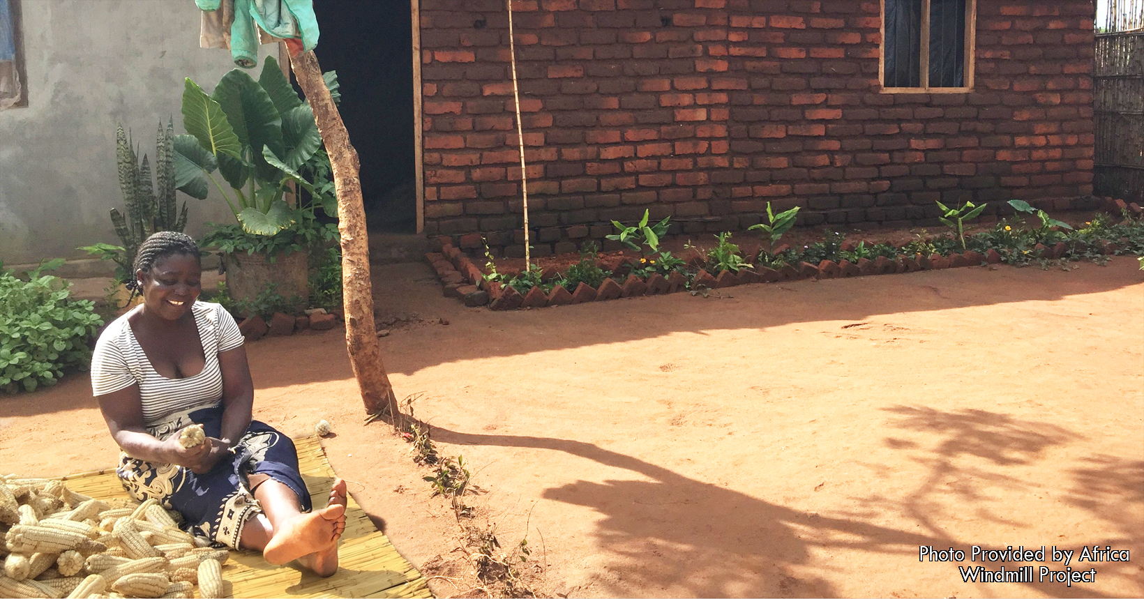 Mercy is sitting in front of her house sitting down with her corn that she recently harvested.